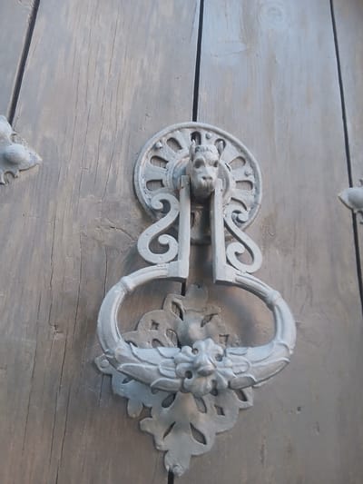 The image shows a close-up of a weathered, ornate metal door knocker attached to a wooden door. The door knocker is made of gray metal and features a design with a lion's head at the top, from which a heavy ring hangs. Below the ring is another decorative element with a stylized face or motif. The wooden door itself is made of vertical planks, showing signs of age and wear with visible grain and cracks. The lighting suggests it is daytime, casting a slight shadow from the knocker onto the door. This door knocker is located on a door within the historic city of Ávila, Spain, a place renowned for its well-preserved medieval walls and architecture.