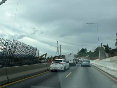 A traffic jam occurs on a cloudy day on a highway in Polichni Municipal Unit, Greece, possibly near Thessaloniki due to the construction. The road is wet, indicating recent rain. The scene is dominated by construction on the left side of the road, featuring a large rebar structure, a yellow excavator, and other construction equipment like cranes. A concrete barrier separates the construction area from the highway. Several vehicles are visible in the traffic jam, including a white Hyundai i30 in the foreground, a white van, a blue Mazda, and other cars further ahead. A tall streetlight stands on the right side of the road, adjacent to a slope with trees and a fence.