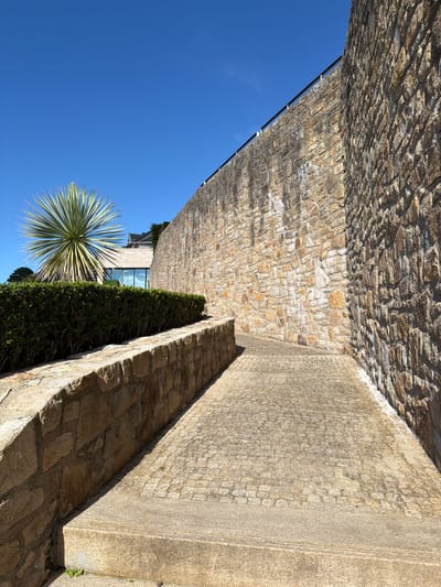 This media file captures an outdoor scene with a strong architectural presence, bathed in bright sunlight. The primary subjects are two contrasting walls, one constructed of rough, uneven stone blocks and the other a smoother, textured concrete. These walls create a narrow pathway or corridor, leading the viewer's eye into the scene.
To the left, a large expanse of clear, vibrant blue sky dominates, suggesting a sunny day. A large, spiky plant, possibly a yucca or agave, is visible against this sky, its green leaves fanning out dramatically. Behind the plant, a modern building with large glass windows can be seen, partially obscured by dense green hedging.
The pathway itself is paved with irregular stone slabs. The stone walls are a warm, earthy tone, with visible mortar between the individual blocks. The concrete wall is lighter in color and has a more uniform, granular texture. Shadows are cast by the walls and the plant, indicating the sun is high in the sky, likely mid-morning or afternoon.
The overall impression is one of a well-maintained outdoor space, possibly part of a private residence, a hotel, or a public garden. The combination of natural elements like the plant and stone with modern architecture suggests a contemporary design. The lighting and clear sky convey a sense of warmth and pleasant weather. There is no visible text in the image. The location context provided is La Clarté, France, which aligns with the Mediterranean-like vegetation and stonework.