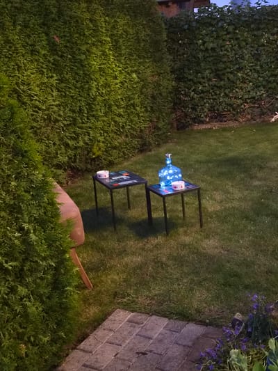 The image shows a garden scene in Bettembourg, Luxembourg, likely at dusk. A lush green hedge forms the backdrop, with a larger coniferous tree to the left partially obscuring the view. In the center of the frame, two small, dark tables are placed on the lawn. The table on the left holds a round white object, possibly a candle or a container, and a decorative item with colorful geometric shapes. The table on the right is adorned with a clear glass bottle filled with blue lights, creating a luminous effect, and another similar white round object. The grass is neatly mowed. In the foreground, a paved walkway made of grey interlocking stones leads into the garden. Small purple flowers are visible at the edge of the walkway on the right. The lighting suggests it's an outdoor evening setting.