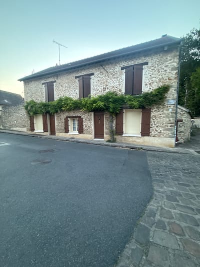 A stone house with dark brown shutters and windows is located on a street in Longpont-sur-Orge, France. The house has a pitched roof and is adorned with climbing green vines. A street sign is visible on the side of the house. The street is paved with asphalt on the left and cobblestones on the right. The sky is clear and light blue. There are no people or animals visible in the image.