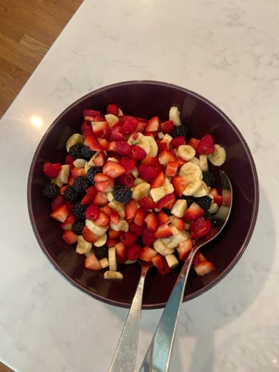 A purple bowl filled with a fruit salad containing sliced strawberries, raspberries, blackberries, and bananas. Two silver serving spoons are on top of the fruit salad. The scene is located inside a kitchen, as indicated by the marble countertop and wooden floor. The coordinates 40.952, -73.708 place the location in Rye, New York, United States. The image is taken from a bird's eye perspective, looking directly down into the bowl. The lighting is bright, creating a slightly reflective surface on the countertop. The image is in focus, capturing the details of the fruit and serving spoons. The background is out of focus, highlighting the bowl of fruit salad. There are no people in the image. The spoons and the fruit salad are the primary subjects. The fruit salad is composed of various fruit slices. The spoons are silver and reflect the light. The bowl is purple with a slight glaze. The background is a white marble countertop with a wooden floor out of focus.