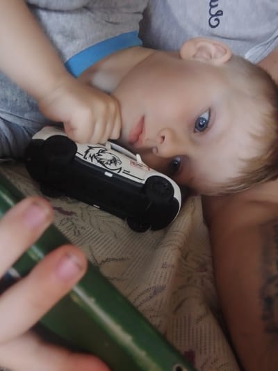 The image shows a close-up of a young child, likely a toddler, lying down with a toy car. The child has fair skin, light brown hair, and striking blue eyes. They are wearing a grey t-shirt with a blue collar and are holding a white toy car with black wheels and a black and white graphic on its side. The child's left hand is resting near their chin, while their right hand is holding the toy car. A portion of a green cylindrical object, possibly a tube or toy, is visible in the lower left corner of the frame, and the child's fingers are touching it. The background appears to be a textured surface, possibly a blanket or bedspread. The lighting suggests an indoor environment. There is no identifiable context to place this image in the city of Pryvilne, Ukraine.
