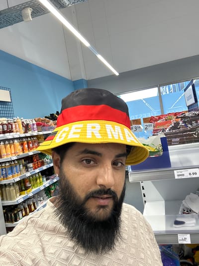 A male individual is depicted in a retail establishment in Pfaffenhofen an der Ilm, Germany. He is shown from the chest up, facing the camera directly. He has a dark beard and is wearing a light-colored, possibly beige, intricately patterned knit sweater. On his head, he wears a bucket hat with a black top band, a red middle band, and a yellow brim. The word "GERMANY" is embroidered in white capital letters on the yellow brim, with "GERMA" fully visible and the subsequent letters partially obscured.
The background reveals a grocery store or supermarket environment. To the left of the individual, multiple shelves are visible, stocked with various bottled condiments such as sauces, mustards, and possibly oils. Price tags on these shelves indicate figures like "3.99" and "1.99." The wall behind these shelves is painted light blue. To the right, shelves display packaged candy and chocolate bars, including brands like Snickers and Reese's. A white price tag on this shelf reads "Schokoladenfußbälle Netz 100g" with a price of "0.99," and another tag shows "1.39." A partially visible sign in the upper right background displays the German word "Kaubonbons." Large windows are present in the far background on the right, showing a reflection of interior text and hints of an outdoor scene. The ceiling is white with a grid pattern, featuring elongated fluorescent light fixtures and visible cable trays. A white speaker is mounted on the ceiling in the upper left portion of the image.