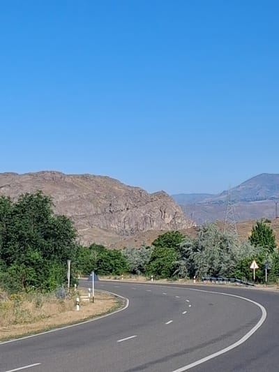 The image depicts a winding asphalt road in the vicinity of Malishka, Armenia. The road curves to the right, bordered by dry, grassy terrain on the left and a mix of green trees and shrubs on the right. In the background, a rugged, rocky mountain range dominates the landscape, with smaller, more distant hills visible further beyond. A few power line towers are also present in the mid-ground. The sky is clear and blue. Road markers and a triangular warning sign are visible along the roadside. There are no people or vehicles present in the image.