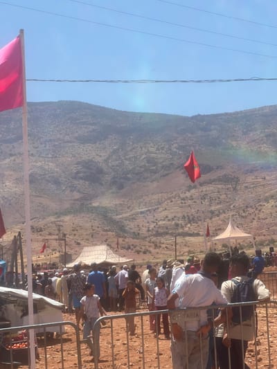 The image depicts an outdoor gathering in Tabia, Morocco. In the foreground, a metal fence separates the viewer from a crowd of people. Behind the fence, several individuals are visible, including children and adults. Some people are interacting with each other, while others are observing the surroundings. To the left, a market stall with tomatoes can be seen. Several tents, possibly for shade or vendors, are set up in the middle ground. Two red flags, one large and one smaller, are prominently displayed on poles. In the background, a mountainous landscape with sparse vegetation dominates the scene under a clear blue sky. Electrical wires are visible overhead. The overall impression is of a community event or market taking place in a rural, arid environment.