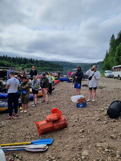 This image depicts a group of people gathered on a rocky shore next to a river in Visherogorsk, Russia. The sky is overcast with scattered clouds. In the background, a dense forest lines the opposite bank of the river. Several inflatable rafts are pulled up onto the shore, suggesting preparations for or the conclusion of a water-based activity, likely rafting. People of various ages are dressed in casual outdoor clothing, some in athletic wear, others in camouflage attire. Some individuals are standing in conversation, while others are observing their surroundings. Objects scattered on the ground include bags, a blue crate, and several paddles lying near the water's edge. In the distance, a white bus and a smaller white van are parked, hinting at transportation support for the group. Two large, orange, netted bundles are stacked on the shore in the foreground, possibly containing gear or supplies.