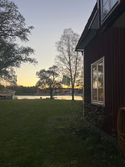 The image depicts an outdoor scene at dusk, consistent with a coastal or lakeside area in Västervik, Sweden. In the foreground, a verdant lawn extends across the lower portion of the frame. To the right, the red, vertically paneled facade of a two-story building is partially visible, featuring a white-framed window on the first floor that reflects warm light from the interior and the sky. A yellow coiled hose hangs against the building's exterior on the far right. Shrubbery grows along the building's base.
The mid-ground features a body of water, reflecting the orange-yellow hues of the setting sun. Along the water's edge, two indistinct individuals are seated on a low embankment. A red bench is situated to their right. To the left, a white, open-sided structure is visible near the water. The distant shore reveals trees, several moored boats, and industrial-type buildings with red roofs, suggesting a marina or port.
Multiple trees are present, including a large, dark-foliaged tree on the far left and a taller, sparser tree in the center-right. A Swedish flag, blue with a yellow Nordic cross, is visible, hanging from a pole or branch in the mid-ground. The sky transitions from a pale blue overhead to a warm orange-yellow gradient near the horizon, indicating late afternoon or early evening light. No active interactions or specific actions are discernible from the two seated individuals.