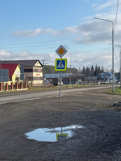 The image shows a street scene in what appears to be Mesyagutovo, Russia. A road runs through the center of the frame, with a puddle reflecting a blue and yellow pedestrian crossing sign on the right side. On the left side of the road, there are two road signs; a yellow diamond-shaped sign and a blue pedestrian crossing sign. In the background, there is a two-story building with a red roof, utility poles with power lines, and trees. The sky is blue with clouds. The overall impression is a somewhat rural or semi-urban environment.