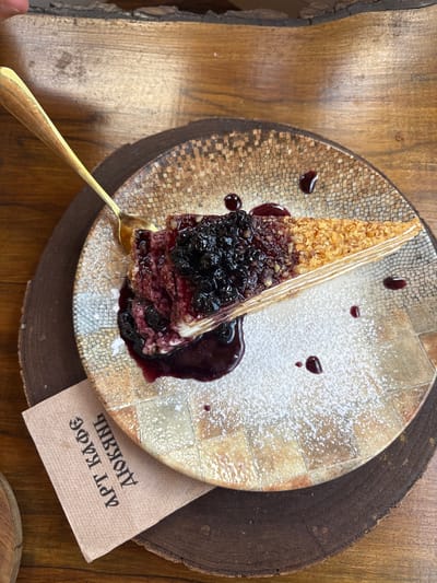 A slice of layered cake, topped with blueberries and a berry sauce, sits on a patterned ceramic plate. The plate is placed on a wooden surface, possibly a table. A gold spoon rests on the plate, partially in the cake. A brown paper napkin with Cyrillic text is tucked under the plate. The scene is indoors, likely a cafe, with natural light illuminating the food. The location is the city of Shiroka Laka, Bulgaria.