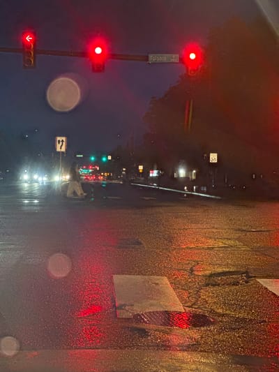 The image depicts a view from inside a car on a rainy night, with the street lights and traffic signals blurred by water droplets on the windshield. On the left side of the road, a traffic light pole has three red lights illuminated: one solid circle and two arrows pointing downward. A street sign attached to the pole reads "Folsom St 2400." This location is Boulder, Colorado, USA. The road surface is wet and reflecting the red lights, along with the headlights of the cars that are further down the street. A speed limit sign is visible with the number 35 displayed. Further down the street are more traffic lights that are green, and red. The scene is dark due to the nighttime setting and rainy conditions.