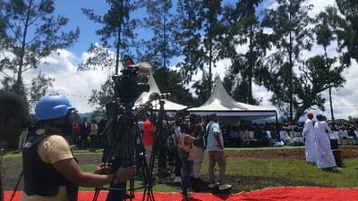 The scene takes place in an open grassy area, possibly a park or a field, in the city of Bukavu, Democratic Republic of Congo, located at the coordinates -1.624 latitude and 29.258 longitude. The area is shaded by a large number of tall trees, with a clear blue sky and white clouds in the background. A crowd of people are gathered in the middle ground, seated on folding chairs under two large, white, tent-like structures. In the foreground, a group of people are focused on filming the event, including a man wearing a blue helmet and a vest, who is operating a camera on a tripod. Another individual, holding a camera with a microphone attached, is also in the foreground. In the far right corner, two individuals dressed in long white robes and a red cap are standing together. There are also several other people, perhaps cameramen or attendees, scattered throughout the scene. The camera is focused on the individuals in the foreground, capturing their actions and the overall event.