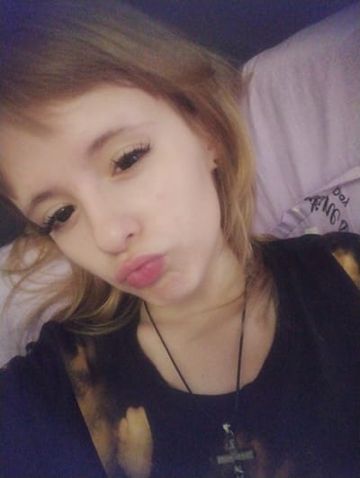The image is a close-up, selfie-style photograph of a young Caucasian female with light brown hair and fair skin. She is making a kissing face. She is wearing a dark-colored shirt with a cross pendant necklace. The background appears to be a bed with a light purple, possibly satin, pillowcase that has some text on it. The lighting is dim and warm. The photograph is not taken in Tashkent, Uzbekistan.