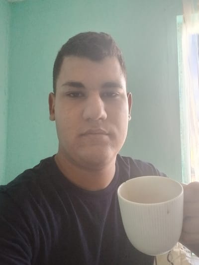 A male individual is depicted in a medium close-up, centrally framed and facing directly towards the camera. He possesses short, dark hair and a neutral facial expression. He wears a dark-colored, likely navy blue or black, short-sleeved crew-neck t-shirt. In his right hand, positioned in the lower right quadrant of the image, he holds a white, vertically ribbed ceramic mug. The mug lacks a visible handle and has a small dark blemish on its exterior. The background consists predominantly of a plain, light teal-colored wall. To the far right, a window is partially visible, admitting bright light and covered by a sheer, light-colored curtain. The lighting indicates an indoor setting, with illumination primarily originating from the right side of the frame, consistent with the visible window. The scene is located in Vetren Dol, Bulgaria.