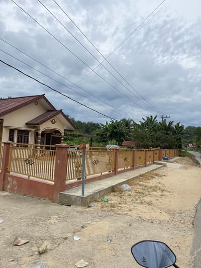 The image depicts a residential street scene in Rantau Pauh, Indonesia. The foreground features a paved surface with scattered debris, including paper and plastic. A motorcycle mirror is visible in the lower right corner, reflecting the overcast sky. A concrete walkway runs along a tan-colored fence with decorative gold metal bars. Behind the fence is a beige house with a dark red roof and brown window frames. A covered porch is supported by columns. Lush green banana trees and other vegetation are prominent in the background. Overhead, power lines crisscross the cloudy sky. The scene appears to be a typical residential area on an overcast day.