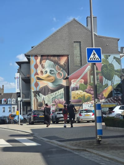 The image captures a street scene in Käerjeng, Luxembourg, on a clear, sunny day. The foreground is dominated by a large mural painted on the side of a building. The mural depicts a stylized duck-like character with rollers in its hair, wearing oversized glasses and holding a newspaper that reads "NEWS FERBER." Various food items, including grapes, cheese, and a jar with a spoon, are also illustrated in the mural.
To the left of the mural, a pedestrian crossing sign is visible. The street itself has a marked crosswalk. Several people are walking on the sidewalk near the mural, and cars are parked or driving on the road. One car has the license plate "KP 9601", and another has "FN 288". A sign for "E.Leclerc" is also visible. The buildings in the background suggest an urban or suburban environment. The overall atmosphere appears to be casual and ordinary, with the mural providing a vibrant artistic element to the everyday scene. The bright sunlight casts long shadows, indicating it's likely midday or early afternoon.