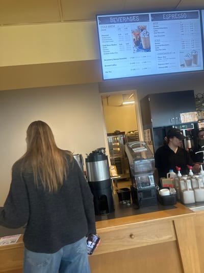 A sideways shot in a coffee shop shows a customer in a gray sweater and jeans looking at her phone. Above her is a coffee brewing station. To the left is a menu board with two sections: "BEVERAGES" and "ESPRESSO". "BEVERAGES" is divided into "COLD BREW" and "Strawberry Banana Smoothie" and "Brewed Coffee" and "Hot Tea". "ESPRESSO" is divided into "HOT", "ICED", and "CUSTOMIZE IT".