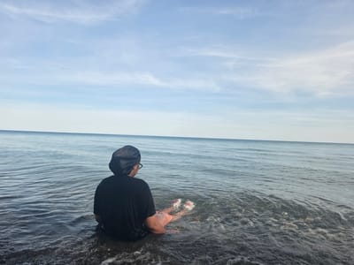 This full-shot image features a person seen from behind, sitting in the shallow waters of a large lake or sea. The individual is wearing a black short-sleeved top and a black satin bonnet or durag, with what appears to be glasses on their face. Their legs are partially submerged in the water, and their feet, adorned with red and white patterned sandals or shoes, are visible just below the surface.

The person is looking out towards the calm, expansive body of water, which stretches to a clear horizon line. The water in the foreground is dark, showing gentle ripples and hints of the bottom, transitioning to a lighter blue further out. Above, the sky is a pale blue with scattered, wispy white clouds, suggesting pleasant and clear daytime weather, likely late morning or afternoon. The setting evokes a sense of peace and contemplation. Given the context of Ajax, Canada, this large body of water is presumably Lake Ontario. There is no visible text in the image.