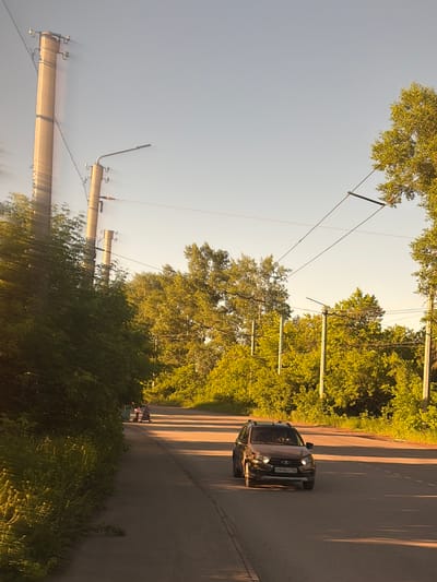 A dark-colored sedan with a license plate that appears to read "0949 AT 177" drives along a paved road lined with lush green trees. The sun is low in the sky, casting long shadows across the road, suggesting it is either early morning or late afternoon. The sky is clear, indicating fair weather. In the distance, silhouetted against the sky, are power lines supported by tall concrete poles. A few people are visible walking on the side of the road, amidst the greenery. The scene is set in a semi-rural or suburban area with a focus on the natural elements of trees and open sky, juxtaposed with the man-made elements of the road and power infrastructure. The overall mood is peaceful and serene, bathed in the warm glow of the setting or rising sun.