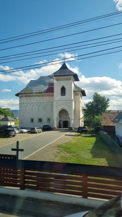 The image depicts the exterior of a religious building, likely a monastery or church, in Vicovu de Sus, Romania. The structure is predominantly white with a dark, steeply pitched roof. A prominent bell tower with a dark spire rises from the front. The building's facade features arched windows and decorative brickwork. A paved walkway leads to the main entrance, which is a large archway. Several cars are parked on the left side of the walkway, and a grassy area with trees is visible to the right. A wooden fence runs across the foreground, partially obscuring the view. The sky is blue with scattered white clouds, and power lines are visible in the upper portion of the image. A cross is cast as a shadow on the walkway in the foreground. The overall scene suggests a sunny day in a rural or semi-rural setting.