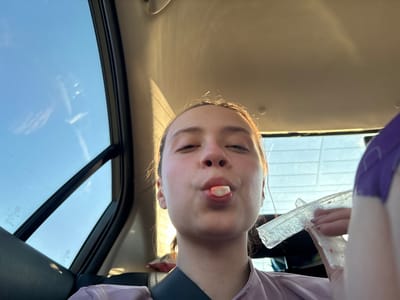 The image shows a person in a vehicle. The person is holding a clear plastic wrapper. There is a piece of candy or food between the person's lips. The image is taken from a low angle, looking up at the person. The background shows a window and the sky outside. The location is unspecified; it is not identifiable as Rakhmatova Polyana, Russia, without additional information.