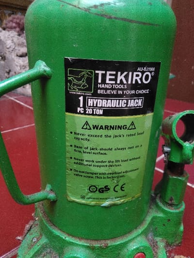 The image displays a used, green, bottle-style hydraulic jack in a close-up, slightly elevated perspective. A black and green label on the jack's cylinder prominently features "TEKIRO® HAND TOOLS," a horse logo, "BELIEVE IN YOUR CHOICE," "1 HYDRAULIC JACK," and "PC 20 TON" with model number "AU-BJ1006." Below this, a "WARNING" section lists operational guidelines such as "Never exceed the jack's rated load capacity" and "Base of jack should always rest on a firm, level surface." The label also includes "GS," "CE," and "JIS JAPANESE INDUSTRY STANDARD" certifications. The jack exhibits a dull green finish with visible wear, dust accumulation, and minor corrosion, especially around its textured base and a robust, curved handle attachment point on its left side. A component of the release valve mechanism is partially visible on the right. The jack rests on a reddish-brown, possibly tiled or paved, surface that shows some dirt. The background is out-of-focus, suggesting a mottled, indeterminate environment. No visual cues within the image indicate its specific location in Mornington, Australia.