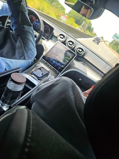 The image depicts the interior of a car driving on a highway, likely near Clamart, France, as suggested by the presence of French road signs. The perspective is from the passenger seat, looking forward. The driver, wearing a blue shirt, is visible with their hands on the steering wheel. The car's dashboard features a large infotainment screen displaying navigation. A glass bottle with a wooden lid is in the center console, next to a smartphone charging on a pad. The passenger in the foreground, partially visible, is wearing a grey top and has red nail polish. The environment outside the car shows a tree-lined highway under a bright, possibly sunny sky.