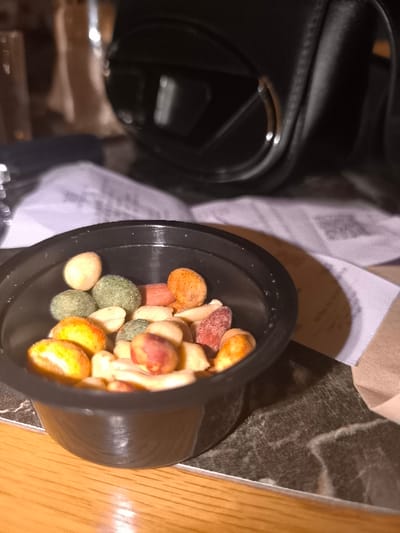 A close-up, eye-level shot prominently features a small, black plastic cup containing a variety of mixed nuts and coated snacks. The nuts display a range of colors, including natural beige peanuts and others coated in vibrant green, orange, red, and pale beige hues. The plastic cup itself is ribbed on its lower exterior and rests on a dark, patterned surface, possibly marble or stone, which in turn partially covers a lighter, wooden table surface visible in the immediate foreground.

The background is softly blurred, providing context without distracting from the main subject. Visible elements include crumpled white papers or napkins, one of which faintly shows a QR code and some unreadable text. A portion of a brown paper bag is also discernible behind the cup. To the upper right, a large, dark, out-of-focus object that appears to be an earcup of over-ear headphones or a camera lens is visible, reflecting ambient light. Another blurry, reflective object, possibly a glass, is present on the far left.

The lighting is warm and artificial, suggesting an indoor setting such as a bar, cafe, or casual dining area, likely during the evening or night. The cup casts a shadow onto the dark table surface. There are no people visible, implying the snacks are prepared for consumption. While the location context is Palaio Faliro, Greece, there are no specific visual cues within the image to confirm this.