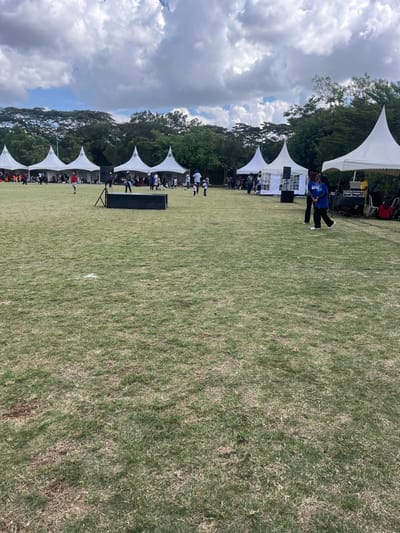 This image captures an outdoor event set on a spacious, well-maintained grassy field in Kalimoni ward, Kenya. A prominent row of white, peaked tents lines the left side of the field, extending towards the background. These tents provide shade for numerous attendees, some of whom are visible seated or standing beneath them, indicating a gathering.

In the mid-ground, two individuals dressed in blue shirts are visible standing on the grass, facing towards the main event area, possibly coordinating or participating in activities. Large, black speaker boxes or stage equipment are positioned on the field in front of the tents, suggesting that a performance, presentation, or public address is taking place.

A lush line of green trees forms a natural boundary and backdrop to the event space. The sky overhead is partially cloudy but bright, indicating fair weather during the daytime. The overall ambiance suggests an organized community event or celebration taking place in a pleasant outdoor setting. No discernible text or specific branding is clearly visible on any of the tents, clothing, or equipment.