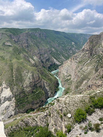 An aerial view of the Sulak Canyon near Chirkey, Russia. A steep-sided gorge contains a turquoise river. The canyon walls are made of layered rock formations with sparse vegetation. A road runs along the edge of the canyon on one side. The sky is partially cloudy.