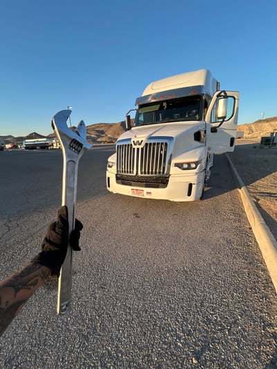 In the city of Salina, United States of America, a close-up shot captures a gloved hand holding a large, silver adjustable wrench. The wrench is positioned in the foreground, with its jaws open. Behind it, a white semi-truck with its driver's side door open is parked on an asphalt surface. The truck has a chrome grille and a license plate that reads "B1 DOBBY". In the background, there are arid hills under a clear blue sky, with other trucks and vehicles visible in the distance, suggesting a truck stop or rest area. The lighting indicates it is either early morning or late afternoon.