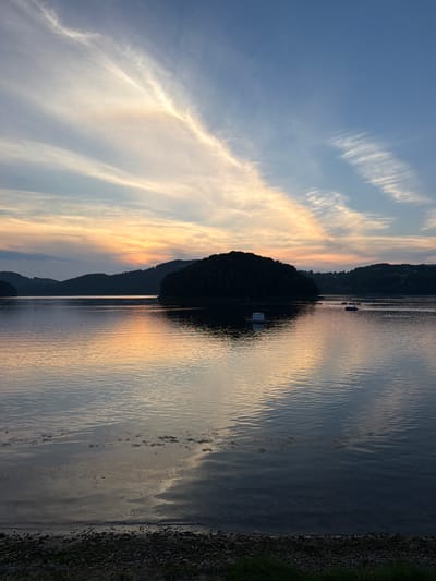 The image captures a serene sunset over a body of water, likely the Gródek Reservoir in Gródek nad Dunajcem, Poland. The sky is painted with wispy clouds, streaked with hues of orange, yellow, and blue, reflecting softly on the calm water's surface. Silhouetted hills, covered in dark vegetation, form the horizon line. In the middle ground, a small, densely wooded island stands out. Several small boats are visible on the water, some with occupants, suggesting recreational activity. A white floating structure, possibly a marker or platform, is present near the island. The foreground shows a pebbly shoreline with some sparse grass. The overall atmosphere is tranquil and picturesque, typical of a peaceful evening by the water.