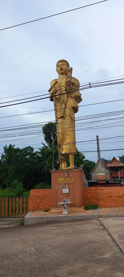The image features a towering golden statue of a monk standing prominently in Ban Thung, Thailand. The statue is detailed and appears to be on a large, decorated base, adorned with elements resembling carved wood and possibly some religious inscriptions. Power lines cut across the view, somewhat obscuring the statue and the sky behind it. In the background, parts of a Thai-style building with ornate roofs are visible amidst the trees. The immediate foreground shows a section of a paved area or road. The overall scene suggests a place of religious significance within a settled or semi-urban environment.