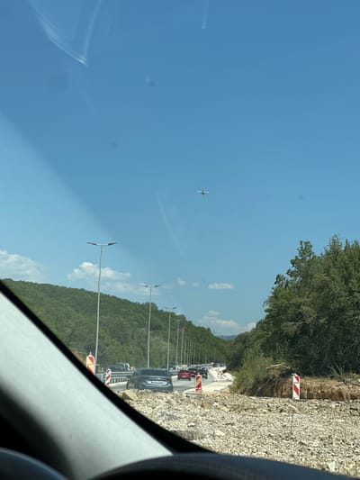The image captures a view from a vehicle driving on a road under construction near Bratešići, Montenegro. In the foreground, a pile of gravel and construction debris is visible on the right side of the road. The road itself is partially paved, with markings indicating ongoing work, and traffic cones with red and white stripes are present. Several cars are driving on the road, some of which are stopped in traffic. In the distance, a line of streetlights stretches along the side of the road. Lush green trees and hills form the backdrop, with a clear blue sky overhead. An airplane is visible in the sky, flying in the distance. The scene suggests a typical day with normal traffic flow, albeit interrupted by road construction.