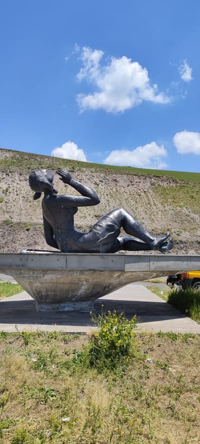 A dark bronze sculpture of a woman in a reclining pose is situated on a concrete platform overlooking a hillside. The sculpture depicts a female figure with her body contorted, knees bent, and arms raised towards her face. She is wearing a skirt or dress. The concrete platform is elevated and has a flared, bowl-like structure beneath it. The background consists of a barren, rocky hillside with sparse vegetation and a clear blue sky with scattered white clouds. In the lower right corner of the image, a portion of a yellow vehicle is visible. The scene appears to be outdoors, possibly in a park or scenic overlook area. The sculpture is located in the city of Nurnus, Armenia.