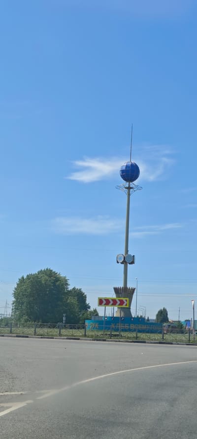 The image shows a tall monument in Buinsk, Russia. The monument consists of a white pillar topped with a blue globe. Below the globe are a series of concentric metal rings. Further down the pillar are two circular, opaque elements. The pillar rests on a decorative base of bundled straw-like structures. At the base of the monument is a blue structure with Cyrillic text that reads "ХЛЕБ ВСЕМУ ГОЛОВА," which translates to "Bread is the head of everything." A yellow traffic sign with red arrows pointing to the right is positioned in front of the blue structure. The surrounding environment includes a road in the foreground, a tree on the left side, and power lines overhead. The sky is blue with a few scattered clouds.