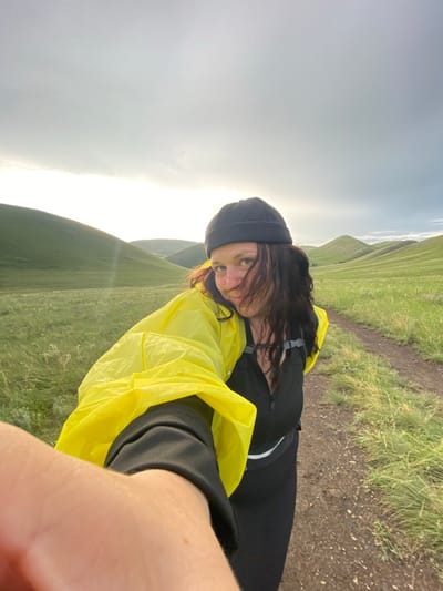 The image shows a young woman wearing a black beanie and a bright yellow hooded jacket over a black zippered vest and black leggings. She has long, dark hair and is wearing a nose ring. She is holding a camera in her left hand, extending it towards the viewer as if taking a selfie. The woman is standing on a dirt path in a vast, grassy landscape with rolling green hills in the background under a cloudy sky. The location is described as the city of Donskoe, Russia.