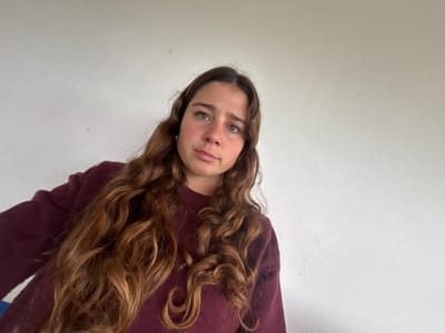 This image, captured in Montemor-o-Novo, Portugal, features a single female individual positioned in the lower-left quadrant of the frame. The subject appears to be an adolescent or young adult with long, wavy, light brown hair flowing over her shoulders and chest. She is wearing a plain, maroon-colored long-sleeved sweater. Her head is slightly tilted, and she is looking directly at the camera with a neutral facial expression. The background consists of a uniformly plain, light-colored wall, likely white or off-white, showing minor blemishes. No other individuals, objects, or specific environmental identifiers are present in the visible scene.