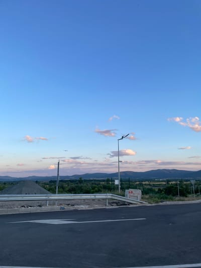 The image depicts a highway under construction or recently completed in the vicinity of Ovoshtnik, Bulgaria. The foreground is dominated by a dark asphalt road with a white directional arrow painted on it. To the left, a pile of gravel is visible, suggesting ongoing construction or maintenance. A metal guardrail separates the road from the embankment. In the middle ground, a light pole with a lamp stands beside the road, and a bird is perched on its arm. Further beyond, a landscape of rolling hills and vegetation stretches towards the horizon, under a vast, pale blue sky with scattered, wispy clouds tinged with pink and orange, indicative of either dawn or dusk. The overall scene suggests a rural or semi-rural setting on the outskirts of a town.