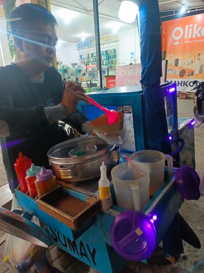 The image depicts a street food vendor in Tenggela, Indonesia, preparing food at a mobile cart. The vendor, a person with dark hair and a black shirt, is actively engaged in their work, holding a pink utensil over a metal cooking vessel. The cart itself is predominantly blue and appears to be a food preparation station, equipped with various containers, including condiment bottles and pitchers. A sign on the cart reads "SUMAY". In the background, a store or market is visible with shelves stocked with items, and a large orange banner with the text "Olike Smart Accessories" is prominent. The scene is illuminated by artificial lights, including purple LEDs on the cart, suggesting it is evening or nighttime. The ground appears to be paved.