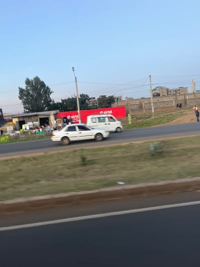 The image depicts a roadside scene in Biashara ward, Kenya, under a clear, light blue sky, possibly during late afternoon or early morning. In the foreground, a dark asphalt road surface is visible, separated by a light brown curb from a grassy median strip containing sparse vegetation.

In the middle ground, a two-lane asphalt road runs horizontally. A white four-door sedan is visible on the left lane, moving from right to left, with slight motion blur on its wheels indicating movement. To its right, a white utility van is positioned on the right lane, also appearing to move in the same direction. A tall, grey street lamp post stands to the left of the van.

On the far side of the road, behind the sedan, multiple makeshift structures, appearing to be small shops or stalls, are constructed from corrugated metal and various materials. Outside these structures, items such as plastic chairs and buckets are arranged. Behind these structures, a large red billboard with "airtel" branding is prominently displayed. A large green tree is visible behind the structures and billboard on the left.

Further right, a dirt shoulder runs alongside the road, leading to a long, light-colored concrete perimeter wall. Beyond this wall, several multi-story buildings, some appearing new or under construction with rooftop water tanks, are visible. Multiple dark power lines traverse the scene above the wall and buildings.

Near the center-right of the wall, two individuals are either seated or standing closely together. Further to the right, a lone individual, dressed in a dark top and light trousers, stands facing right, holding a red and white object. Their actions appear to be stationary observation or waiting. No direct interactions between individuals are discernible.