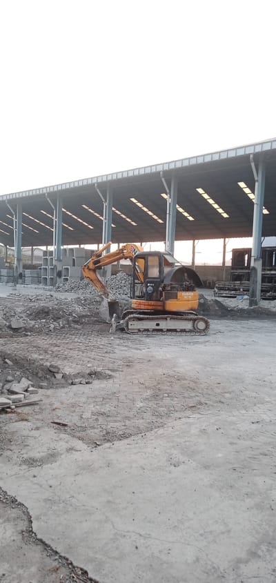 The image shows a yellow Komatsu PC75 excavator in an outdoor industrial setting in Sumput, Indonesia. The ground is a mix of concrete and dirt, littered with concrete debris. Behind the excavator, there is a pile of concrete blocks and building materials. A large open-sided structure with a corrugated metal roof supported by steel beams is in the background. The sky is overcast. There are no people visible in the image.