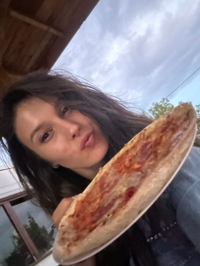 A young woman with long, dark hair is taking a selfie outdoors, holding a slice of pizza up to the camera. She is making a kissy face. The woman is wearing a denim jacket. In the background, there is a wooden overhang and a cloudy sky. The image appears to have been taken in Golyam Chardak, Bulgaria.