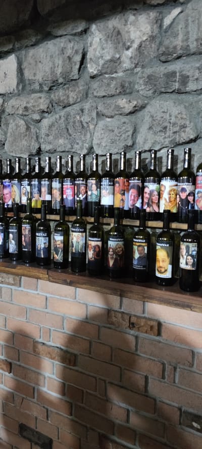 In Voskevaz, Armenia, a row of wine bottles is displayed on wooden shelves against a stone wall. The bottles have labels featuring photographs of people. The top shelf holds a longer line of bottles, and the shelf below holds a shorter line. The bottles are arranged in a neat, organized manner, suggesting a display or a collection. The background stone wall adds a rustic and possibly historical ambiance to the scene. The overall impression is one of a winery or a wine shop in Voskevaz.