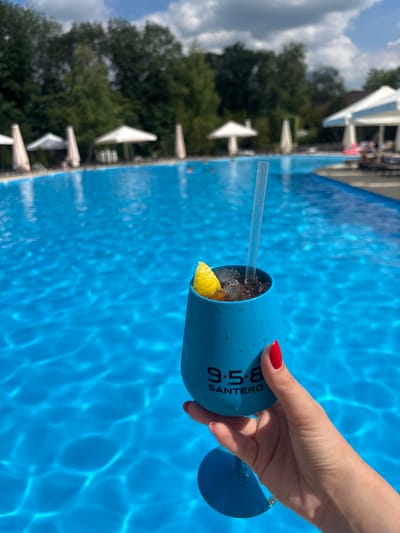 A person's hand with bright red nail polish holds a bright blue tumbler with a clear plastic straw. The tumbler contains a dark beverage with ice cubes and a slice of lemon. The tumbler has "9.5.8 SANTERO" printed in white text. The hand and tumbler are positioned over a bright blue swimming pool with visible water ripples. In the background, there are trees, white umbrellas, and what appears to be a building. The scene suggests a sunny day, likely outdoors at a resort or pool area. The image is oriented vertically, with the top of the frame showing the sky and clouds, and the bottom showing the pool's surface.