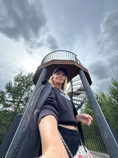 A woman, wearing a black baseball cap, black zip-up jacket, black sweatshirt, and black sweatpants, stands on a wooden platform of an observation tower. She is holding her phone to take a selfie. The tower has a spiral staircase on its right side. Pine trees surround the tower. The sky is overcast with grey clouds.