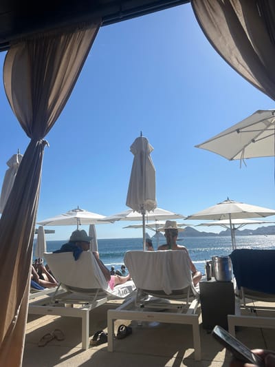 The image shows a beach scene in Cabo San Lucas, Mexico. Several people are relaxing in white lounge chairs under white umbrellas. The ocean is visible in the background with some land. There are brown curtains hanging from the sides, suggesting a cabana-like structure. Some personal belongings, such as sunglasses and sandals, are scattered around the lounge chairs. The sky is clear and blue.