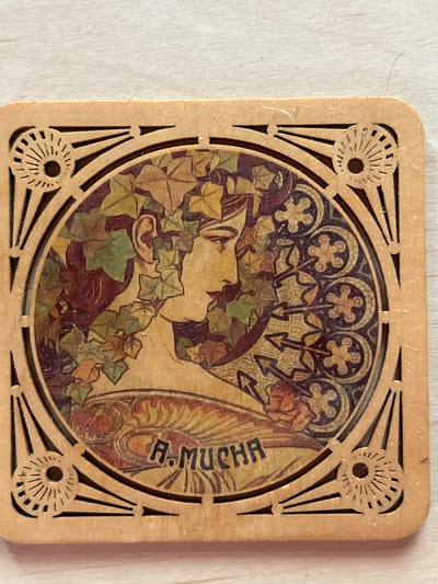 A square wooden coaster, approximately 4 inches by 4 inches, features a decorative inlay in the style of Alphonse Mucha. The central image depicts a woman's profile, adorned with leaves and ivy. Her hair is dark, and she has a delicate facial structure. The inlay is detailed with floral and geometric patterns surrounding the central portrait. The artist's signature, "A. MUCHA," is visible in black lettering at the bottom of the central image. The coaster has a wooden border with decorative cut-out elements, including radiating lines and circular motifs with spokes. The surface of the coaster appears slightly worn. The background of the image is a light-colored, textured surface, likely a table. The scene is lit from above, casting subtle shadows. The object is entirely indoors, and no specific city or geographical location is identifiable from the image itself.