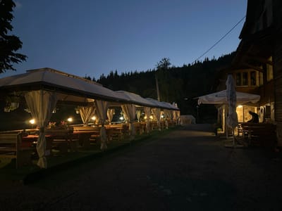 The image depicts an outdoor dining area at dusk in Bogoslov, Bulgaria. A covered seating area with white curtains and multiple tables and benches stretches across the foreground. Each table is set with red and white checkered cloths and illuminated by warm, bright lights, suggesting a welcoming atmosphere. Flower pots with pink flowers are visible on some tables, adding a touch of color.

To the right of the covered seating area, a rustic wooden building with windows and a balcony is visible. A large white umbrella stands in front of the building, partially obscuring it. In the background, a dense forest covers a hillside, silhouetted against a deep blue twilight sky. A single faint star is visible in the sky.

A lone figure is seated at a table on the balcony of the wooden building, their back to the camera. The figure is wearing dark clothing and appears to be looking down, engrossed in something. The overall mood is peaceful and serene, with the lights of the dining area creating a warm contrast against the darkening landscape. The time of day is clearly evening or twilight, as indicated by the deep blue sky and artificial lighting.

On the umbrella, partially obscured text in Cyrillic script is visible, though it is difficult to read clearly. The scene suggests a restaurant or guesthouse providing outdoor dining services in a natural, wooded setting.
