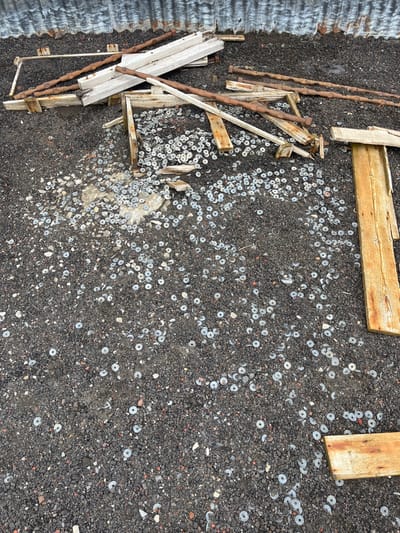 A ground covered with dark, coarse gravel is littered with numerous metal washers. Scattered amongst the washers are several pieces of weathered wood, some forming makeshift structures, along with rusty metal bars. The scene suggests a derelict or abandoned location, possibly a former construction or industrial site. A corrugated metal sheet is partially visible in the image's corner.